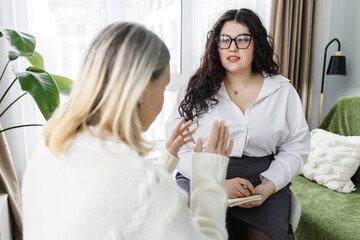 Two people talking in cabinet, young woman therapist listening to complaints and issues of her female client, gesticulating while explaining troubles and confused situation of her life
