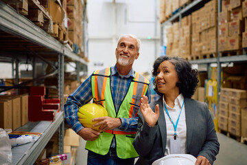 Happy female inspector and manual worker talking while walking through distribution warehouse.