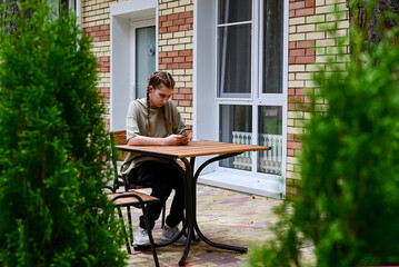 Young person sits alone at a table using a smartphone in an outdoor setting surrounded by greenery and a brick building