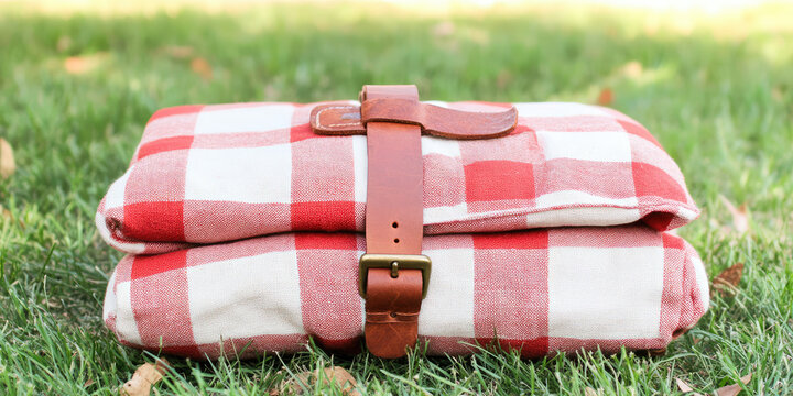 Folded red and white checkered blanket with a leather strap sits on green grass. Cozy, outdoor, picnic, leisure