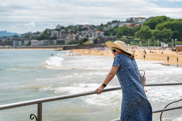 Woman wearing hat and dress looking at the sea views next to Ondarreta beach, San Sebastian, Spain.