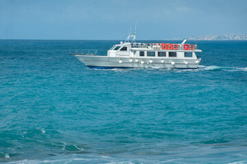 Passenger excursion boat in the turquoise waters of the Mediterranean Sea and city silhouettes in the distance, Italy