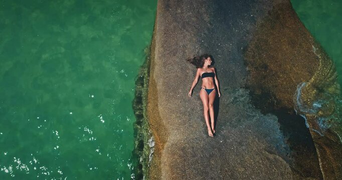 A person relaxes on a large, sunlit rock by the shoreline, surrounded by clear turquoise water. The tranquil scene captures the essence of a perfect day outdoors.