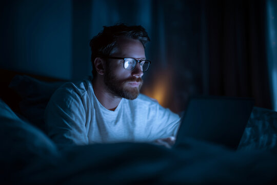 A man is sitting on a bed with a laptop in front of him. He is wearing glasses and has a beard. Concept of relaxation and leisure, as the man is using his laptop in a comfortable setting