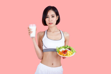 Healthy Asian woman in fitness outfit holding a glass of milk and a fresh vegetable salad. Concept of clean eating, nutrition, and wellness on pink background.