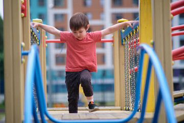 A young boy carefully walking on a playground bridge, holding onto the railings for balance. Child...