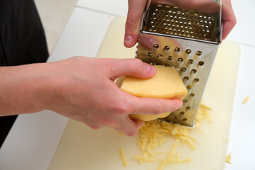 Close-up of hands grating a block of hard cheese using a stainless steel box grater on a kitchen counter.