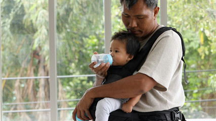 Father feeding baby from bottle while holding them in chest carrier, showing love and parenting care in bright room with nature background