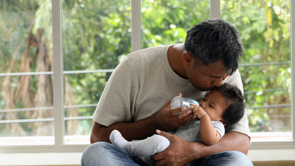 Father lovingly feeding baby with milk bottle while sitting on floor, gently kissing baby head,...
