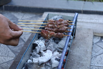 A woman's hand holds a skewer. Goat satay is being grilled over hot coals. Fresh meat is being grilled using a wooden skewer over hot coals. Side view photo.