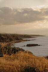 Coastal landscape bathed in golden sunset light, with dramatic cliffs and calm sea waves stretching into the horizon. Peaceful and timeless Mediterranean atmosphere.