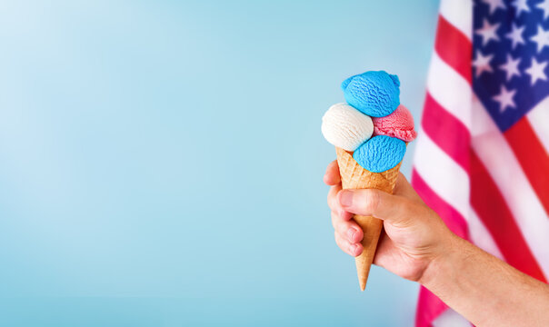 4th of July Holiday background. Mans hand holds colorful ice cream cone with red, white and blue scoops patriotic colors on blue background and American flag. Happy Independence Day concept. Mock up.