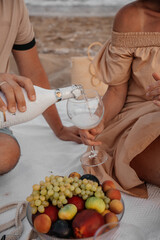 Romantic seaside moments. A couple holds hands near the ocean and enjoys a cozy beach picnic with fresh fruits and a sparkling drink at sunset.