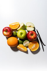 Top-down view of fresh fruits including apples, oranges, kaffir limes, and vanilla pods on white background. Bright natural light creates strong shadows, emphasizing texture and color contrast.