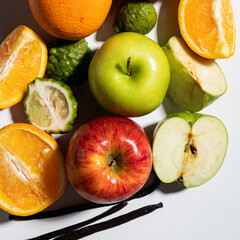 Flat lay of whole and cut apples, oranges, kaffir limes, and vanilla pods on white background with shadows