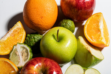Close-up of sliced and whole apples, oranges, and kaffir limes arranged on white background with natural shadows