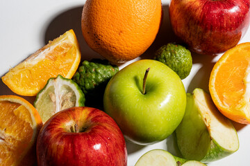 Assortment of fresh fruits including green apples, red apples, oranges, and kaffir limes arranged on a white background with strong shadows and bright lighting emphasizing texture and freshness.