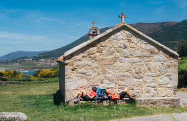 Couple of pilgrims with backpacks lying and resting on bench next to old church on Camino...