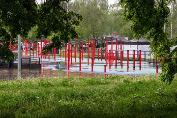 Sports ground with red exercise machines outdoors on green lawn. Green trees around