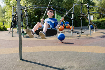 An 11 year old boy wearing glasses and a blue T-shirt is riding a swing in a park on a playground