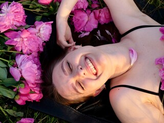 Happy smiling young woman in summer outdoors in a field with pink peony flowers enjoying life in a...