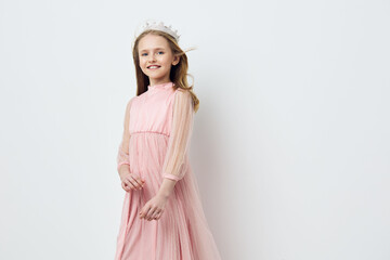 Smiling girl in a pink dress and crown, exuding joy and innocence against a soft white background,...