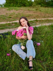Fototapeta premium Happy smiling young woman in summer outdoors in a field with pink peony flowers enjoying life in a moment at sunset