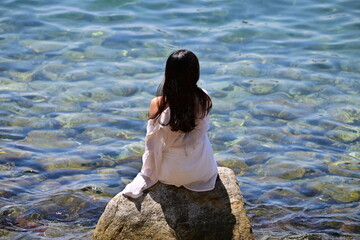 A young, Vietnamese Woman sits o n rock nearby the sea while she looks to the incoming ships.