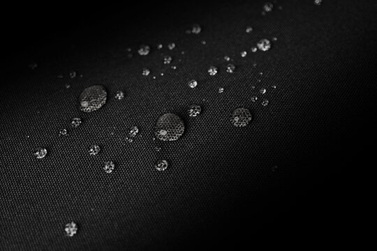 Macro photo of water droplets on black waterproof fabric, showing hydrophobic properties with a clear grid texture under soft lighting, highlighting resistance and surface detail.