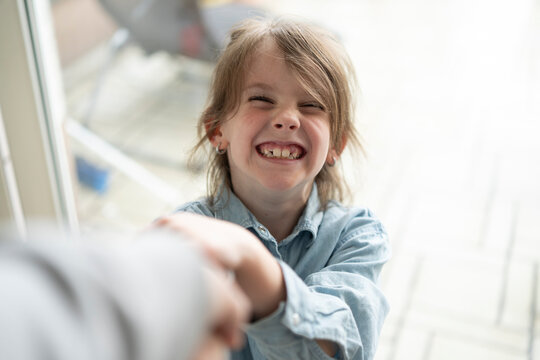 Cute girl in denim shirt pulls parent's hand with mouth open and teeth bared against blurred light window. Dental emotional children concept