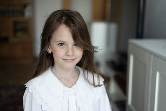 Photo of a beautiful smiling little girl in a white blouse in a room with a blurred background. Concept of beauty and children's emotions