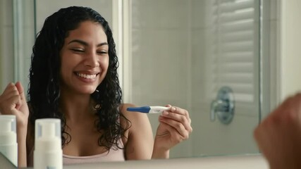 Happy Smiling Hispanic Woman Celebrating Positive Pregnancy Test Result with Fist Pump Looking in Bathroom Mirror, Parenthood Journey Beginning Concept