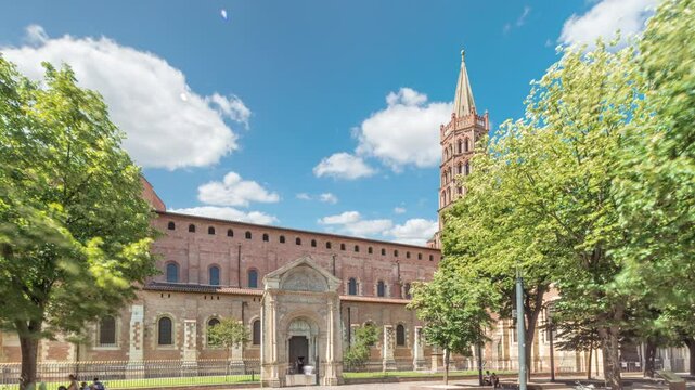 The cobbled parvis of the Basilica of Saint Sernin in Toulouse, France, timelapse hyperlapse. Largest romanesque church in Europe with brick bell tower, arched doors, green trees and blue cloudy sky
