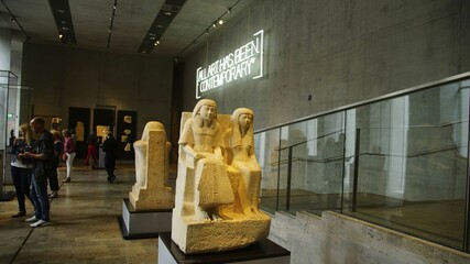 Museum visitors observing limestone statue depicting pharaoh amenemhat iii and spouse within modern art gallery interior