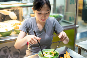 Asian woman enjoying Phở with spring rolls at a bustling street food stall in the evening