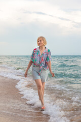 Happy adult blonde woman walking along the seashore on a cloudy summer day. Vacation at sea.