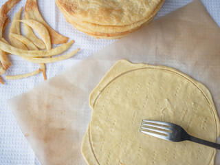 Fork piercing a raw layer rolled out on a baking paper, a pile of baked thin buttery layeres, and dough scraps in the background ready to make a shortcrust puff pastry Napoleon cake.