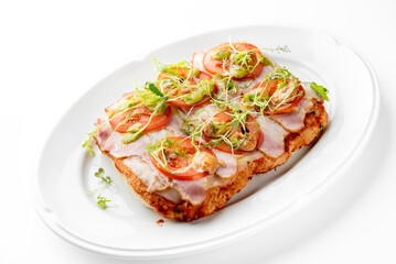 An open-faced sandwich with sliced ham, tomato, and microgreens, drizzled with green sauce, served on a white plate. Fresh ingredients create a colorful and appetizing presentation