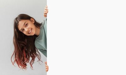 Happy Preteen Girl Posing Hiding Behind Empty White Board And Smiling To Camera In Studio Over Gray Background. Child Holding Paper Poster Advertising Your Text. Panorama, Mockup