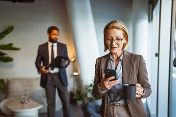 mature businesswoman use mobile phone and drink coffee at workplace