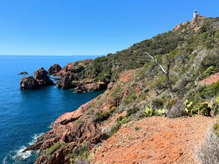 Coastal landscape in the Esterel region of southern France with dramatic red cliffs, deep blue sea, rugged rocks, and a distinctive observation tower perched on the hilltop.

