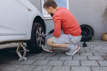 Close-up of a man changing a car tire in front of his house. The vehicle is lifted with a jack,...