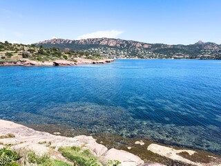 Clear coastal view of the Esterel mountains and turquoise Mediterranean Sea in southern France, with rocky shoreline and transparent water under a bright blue sky.

