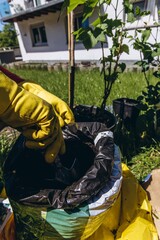 Man wearing yellow rubber gloves plants green currant bush into pot with metal garden trowel on a sunny summer day outdoor gardening concept