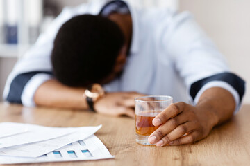 Difficulties at job, bankruptcy concept. Drunk young african american businessman laying on workdesk with documents, drinking cause of stress, holding glass of whiskey, office interior, copy space