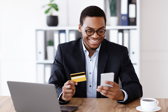 Online shopping, e-payment service concept. Happy african american businessman holding credit card and mobile phone, making e-payments while working with laptop in his office, free space