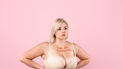 Body Acceptance. You are beautiful motivational inscription on chest of plus size woman, curvy young lady in bra posing over beige studio background, keeping hands on waist and looking away
