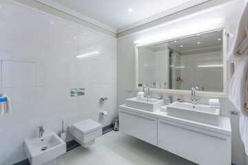 Luxurious bright bathroom, decorated with white tiles, with a large mirror over the double sink. Bidet and toilet.