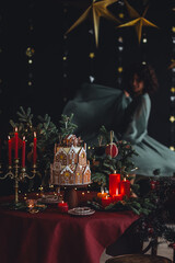 Beautiful woman in knitted sweater making handmade homemade delicious gingerbread house for family Christmas celebration. Elegant table setting. Cozy home atmosphere, festive mood, candlelight