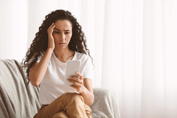 Shocked young woman looking at smartphone screen, sitting on sofa at home, got bad news, copy...